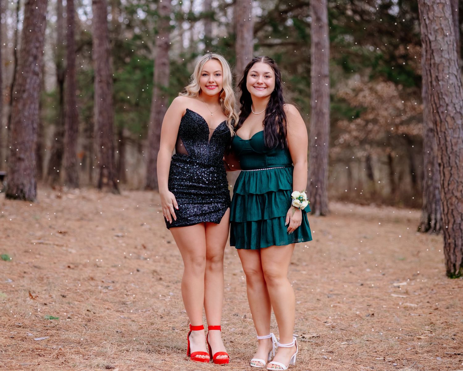 Two people in festive dresses stand together on pine needle covered ground in a forest setting.