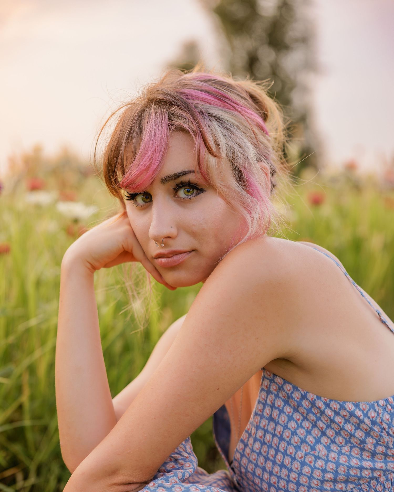 Young woman with pink-highlighted updo hair sitting in a wildflower field wearing a floral dress at sunset.