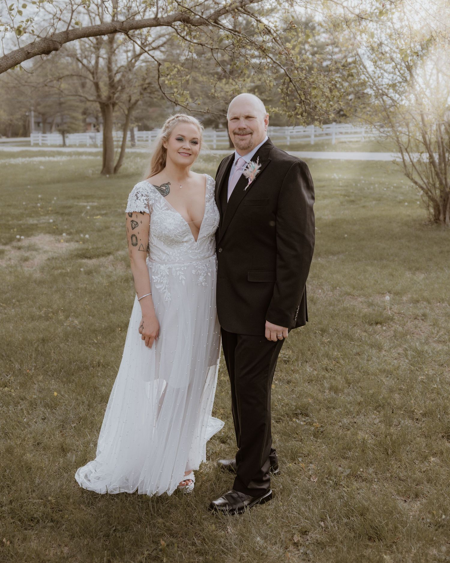Bride in white lace wedding gown and groom in black tuxedo posing together outdoors on green lawn.