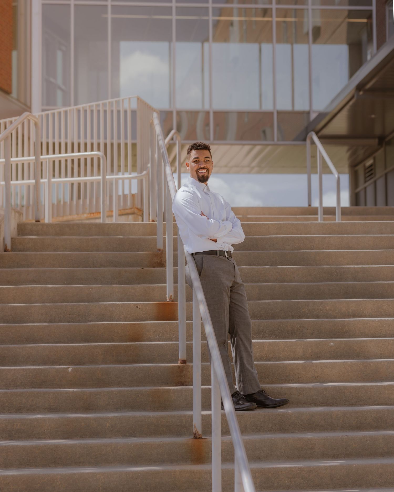Young man in white shirt and gray trousers stands with arms crossed on modern building staircase.