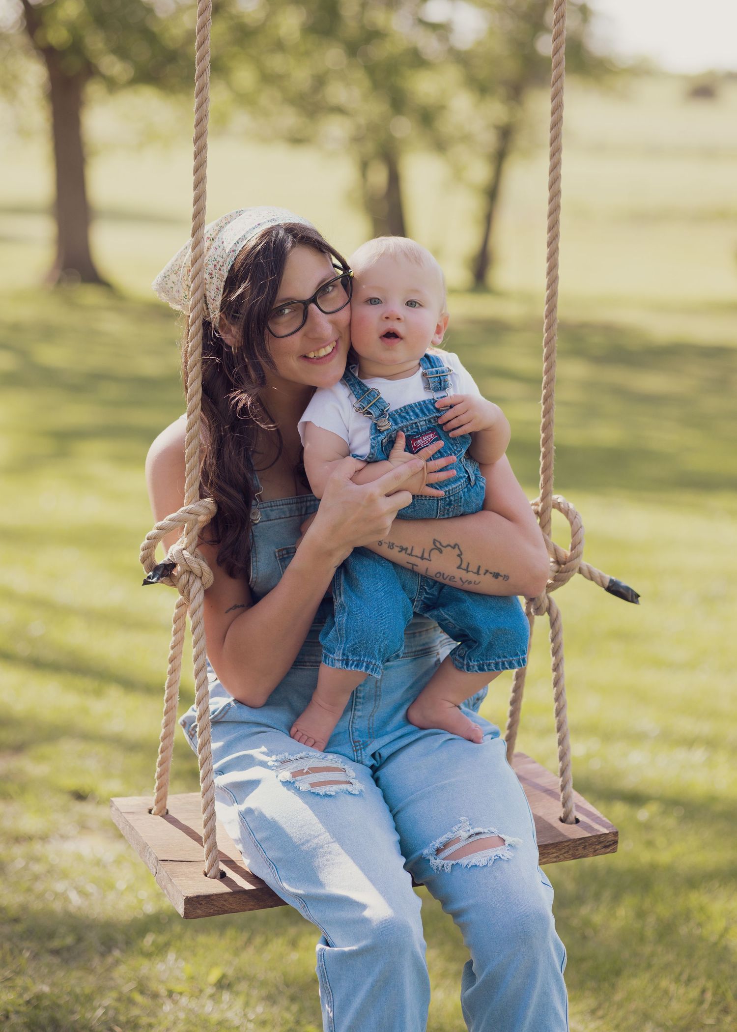 A young woman wearing glasses and overalls sits on a rope swing holding a baby in denim overalls outdoors.
