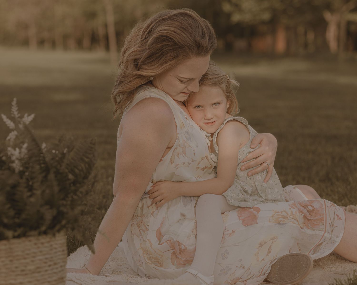 Mother tenderly embracing young daughter in matching floral dresses during golden hour outdoor photo session.