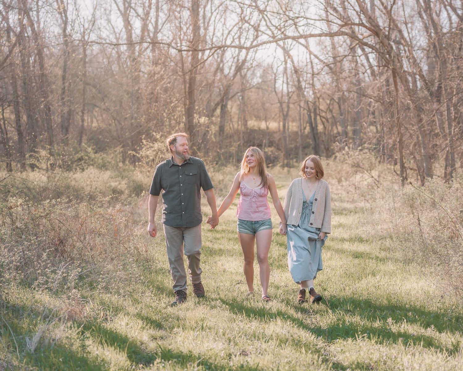 Three people walking hand-in-hand through a sunlit meadow surrounded by bare trees in a peaceful outdoor setting.