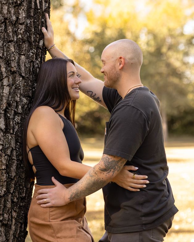 A smiling couple embraces near a tree in a sunlit park, the woman in a black top and tan skirt, the man with tattoos.