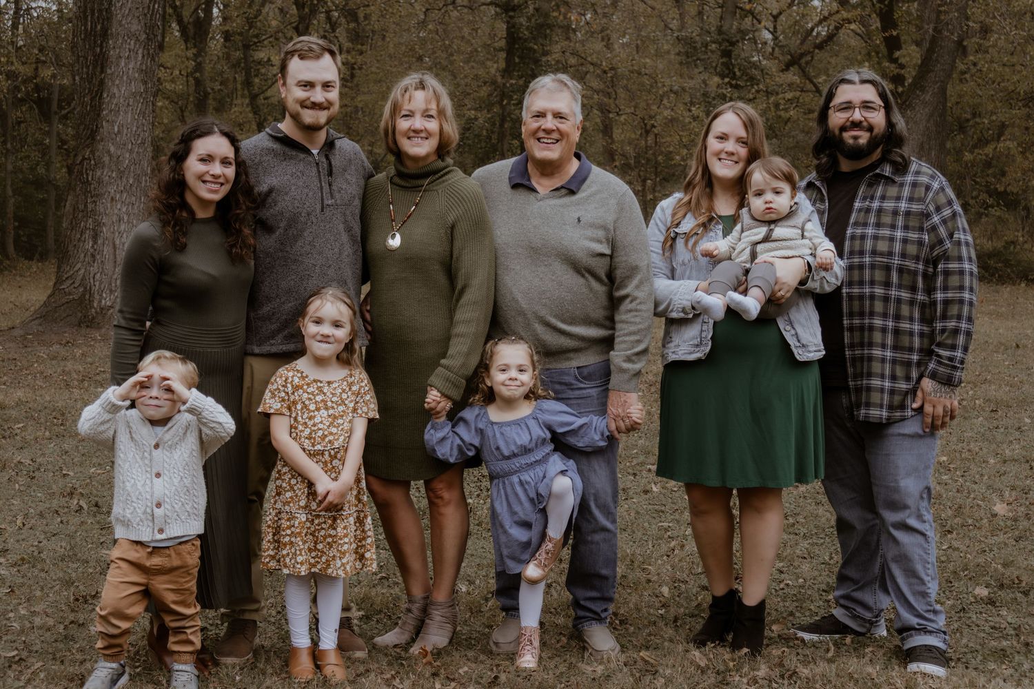 Multi-generational family portrait outdoors in autumn woods, smiling group of adults and young children posing together.