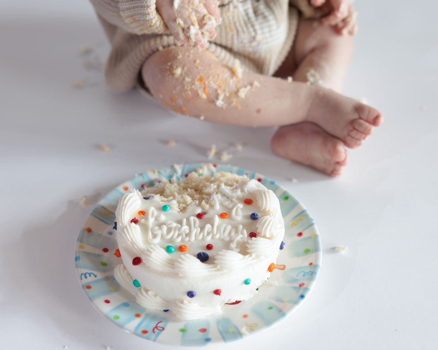 Baby smashing a white frosted birthday cake with colorful sprinkles on a decorative paper plate.
