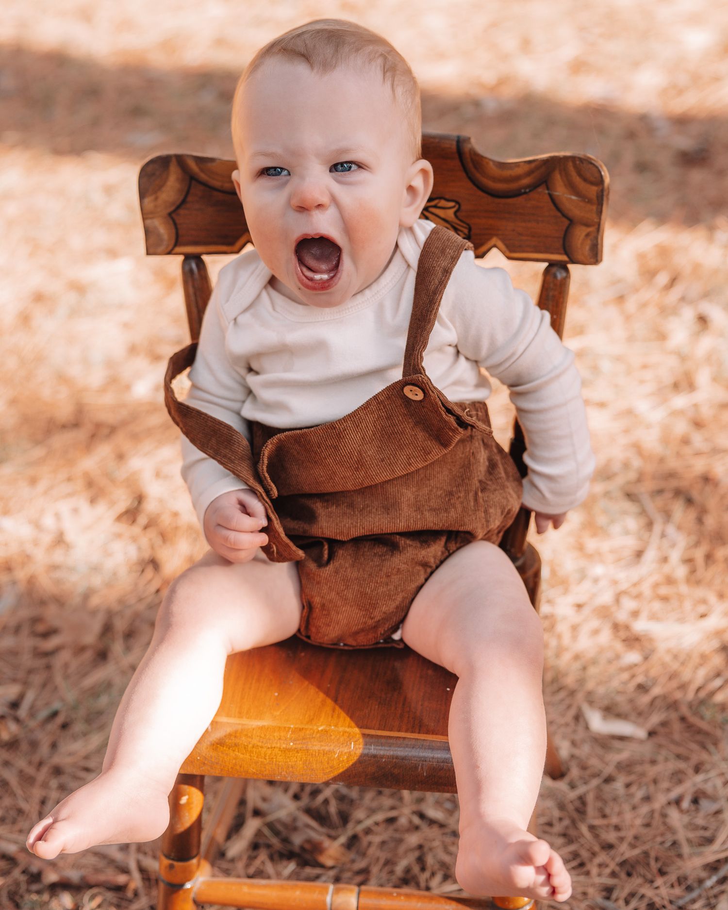 A baby wearing a brown ruffle dress and yellow shorts smiles and plays while sitting on a wooden chair.