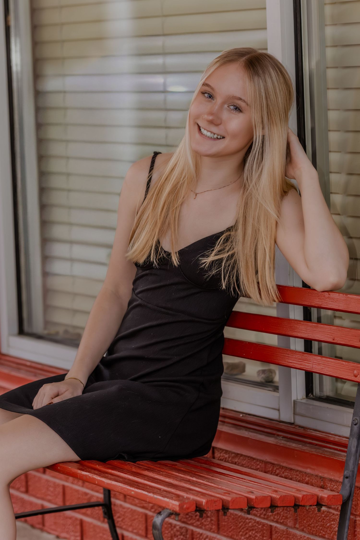 A person in a black dress sits smiling on a red bench outside a building with window blinds visible.
