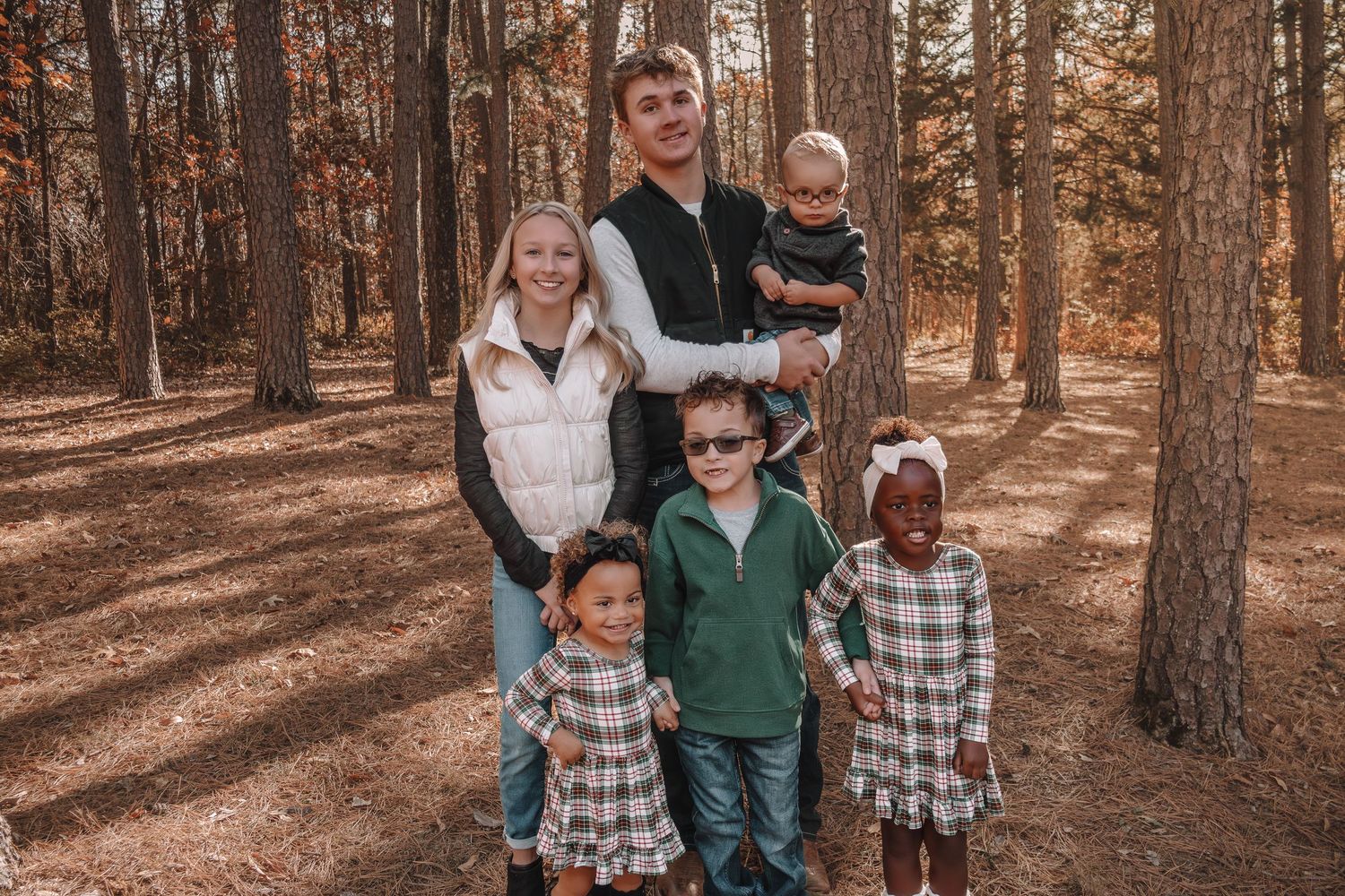 A family wearing coordinated green and plaid outfits poses together in a sunlit autumn forest setting.