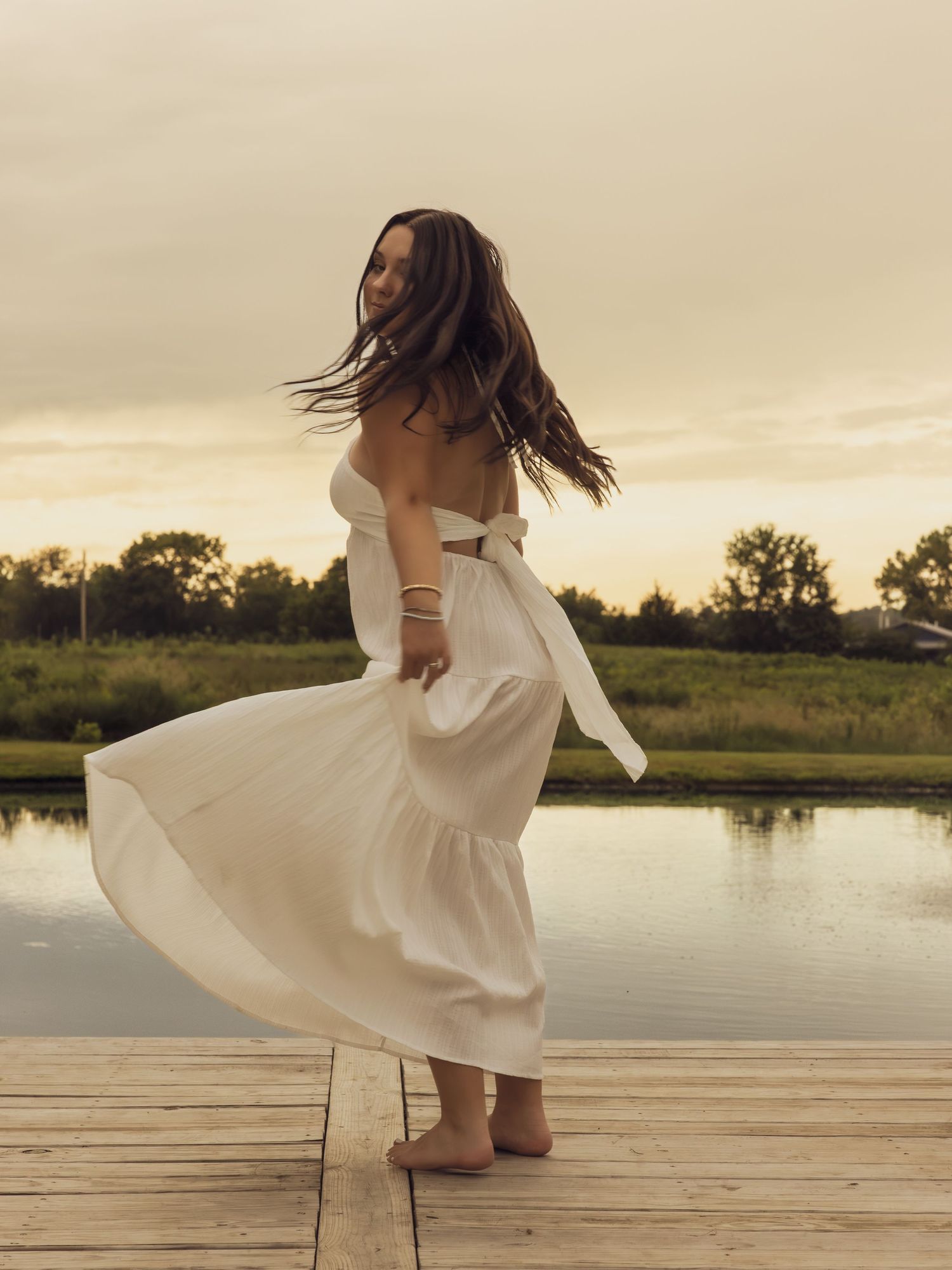Brunette woman twirling in white dress on wooden dock at sunset by a calm lake.