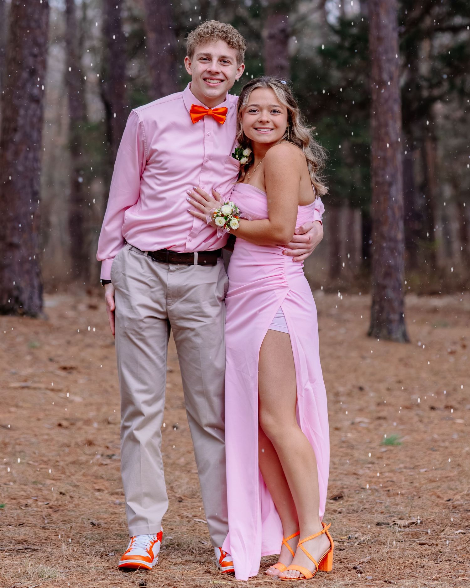 A couple in formal attire with pink and khaki outfits pose together in a wooded outdoor setting during a special occasion.