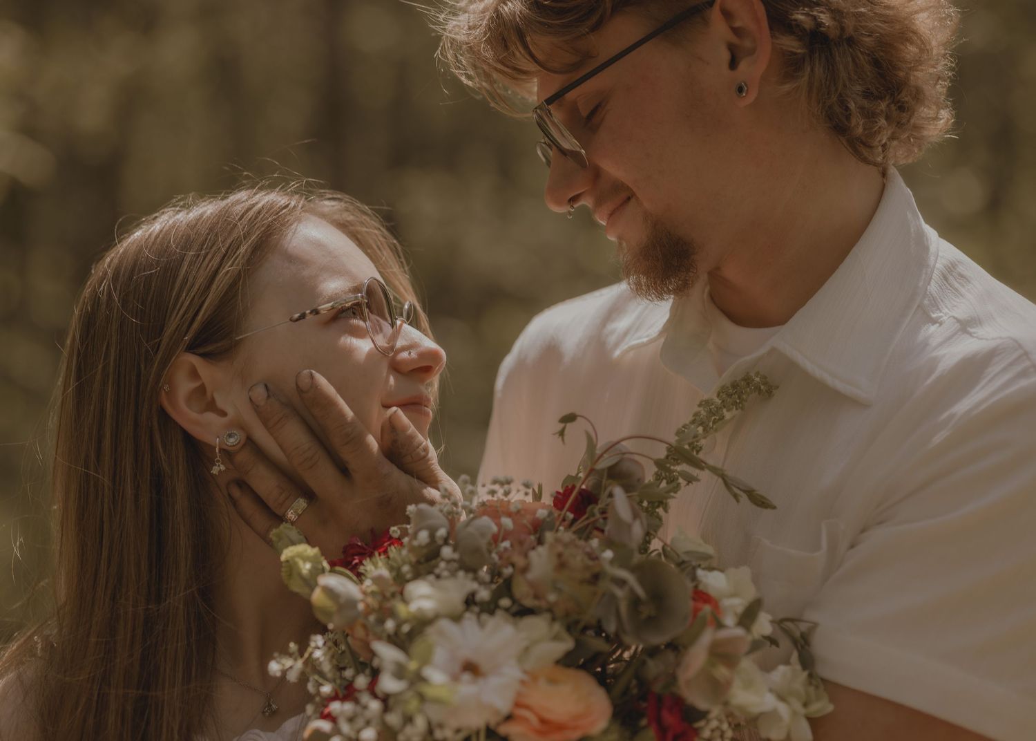 A couple shares a tender moment, the woman holding a colorful bouquet in a sunlit forest setting.