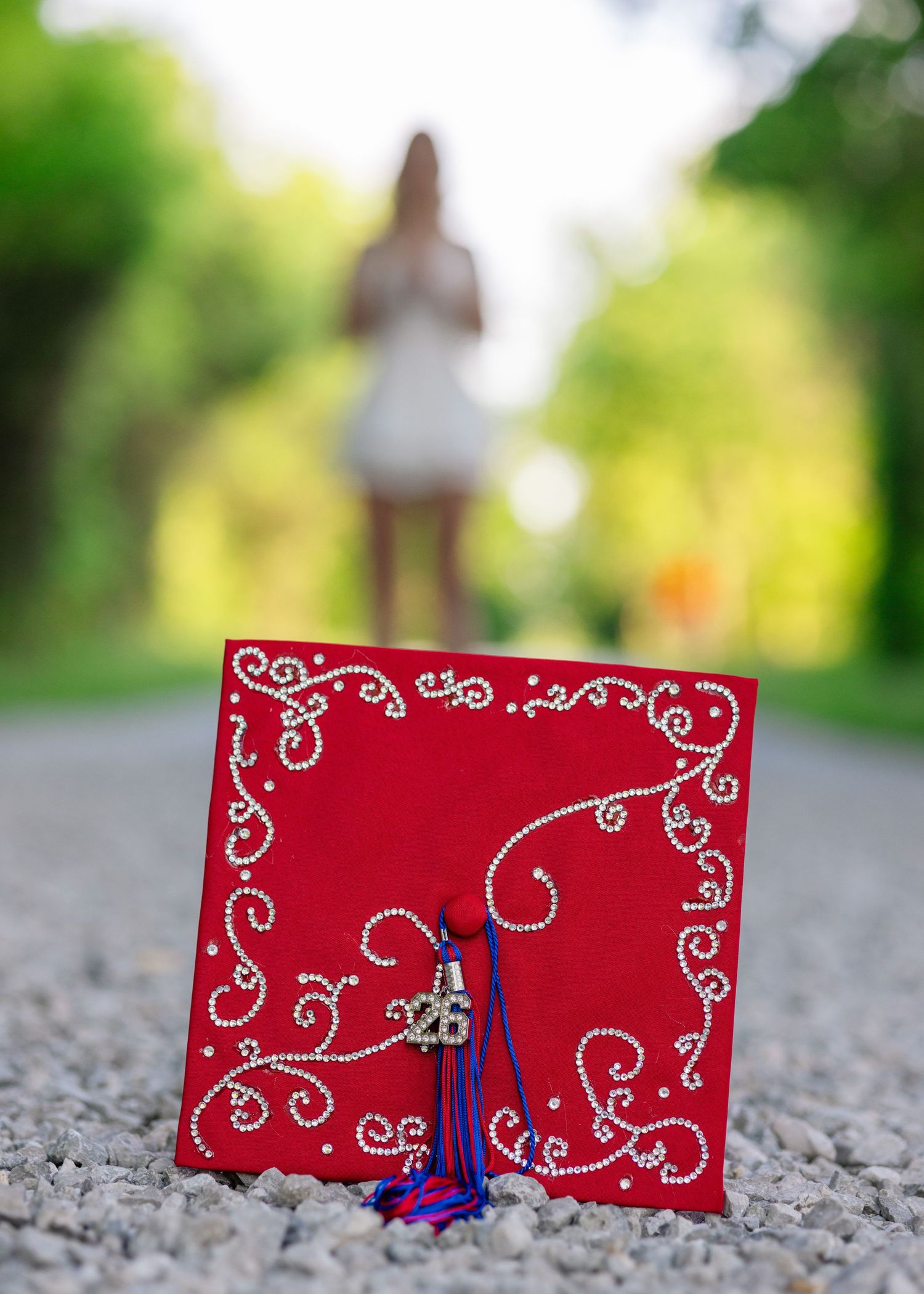 Red decorated graduation cap with rhinestone swirls and blue tassel on gravel path, blurred graduate in white dress behind.