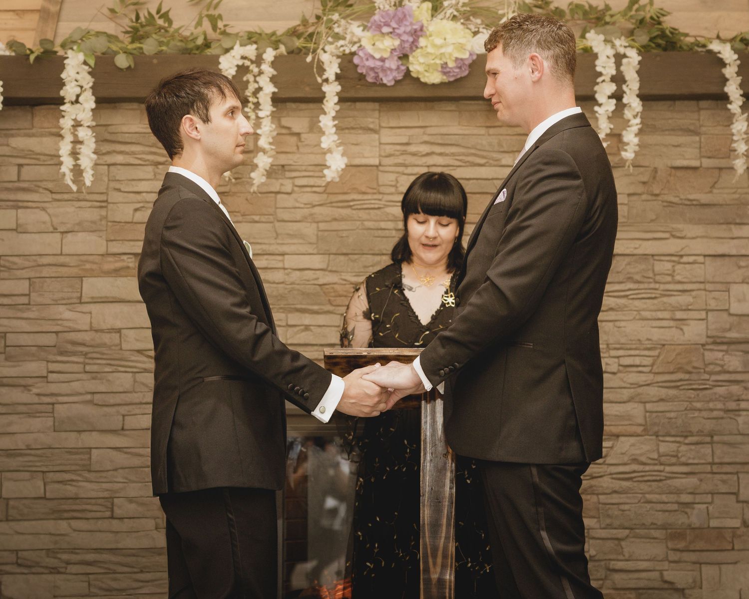Two grooms hold hands during an intimate wedding ceremony officiated by a woman before a stone fireplace.