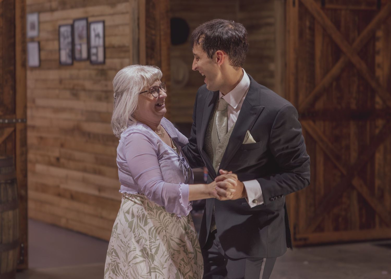 A young man in a suit dances with an older woman in a floral skirt inside a rustic barn venue.