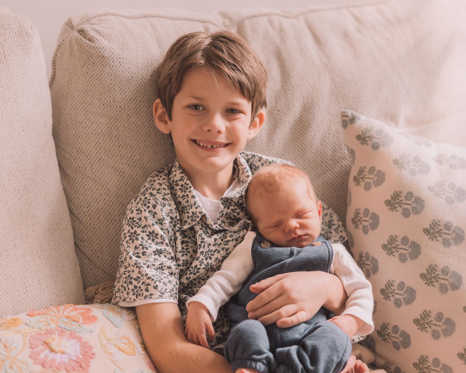 Young boy smiling while gently holding a sleeping newborn baby on a couch with floral pillows.
