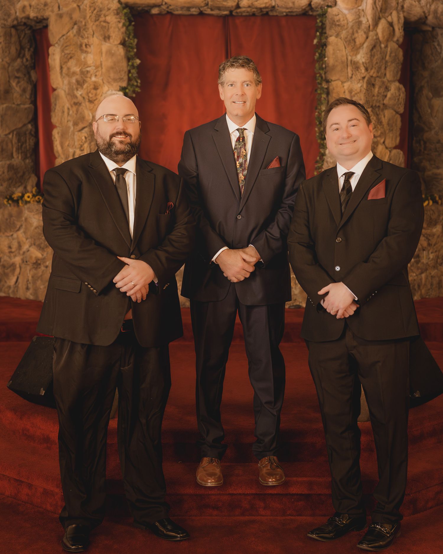 A group of men in dark suits stand together in a formal setting with stone walls and red carpeting.