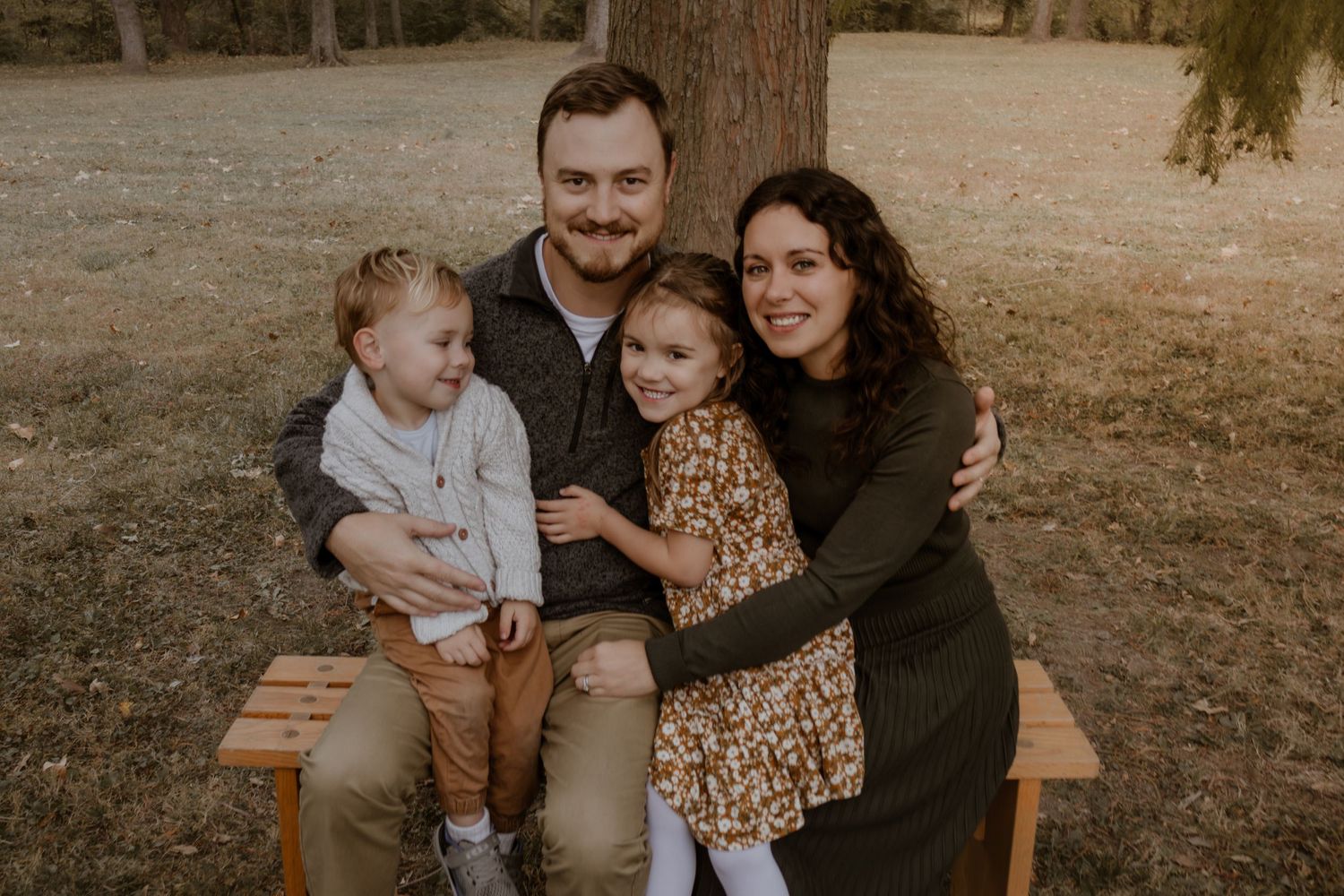 A family sits together on a wooden bench in a park during golden hour, creating a warm and intimate outdoor portrait.