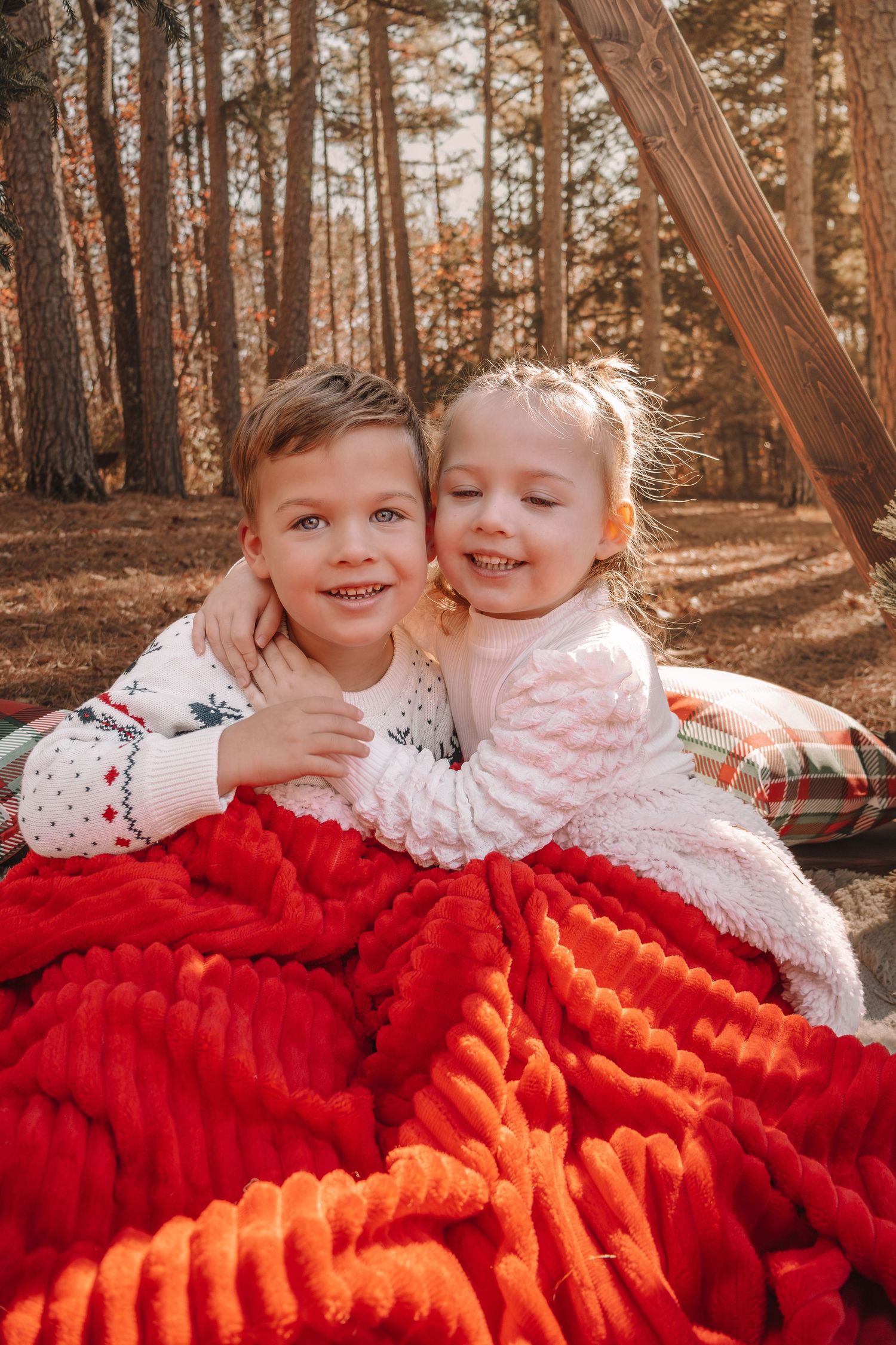 Two children snuggle together on a red and orange blanket in a sunlit autumn forest setting.