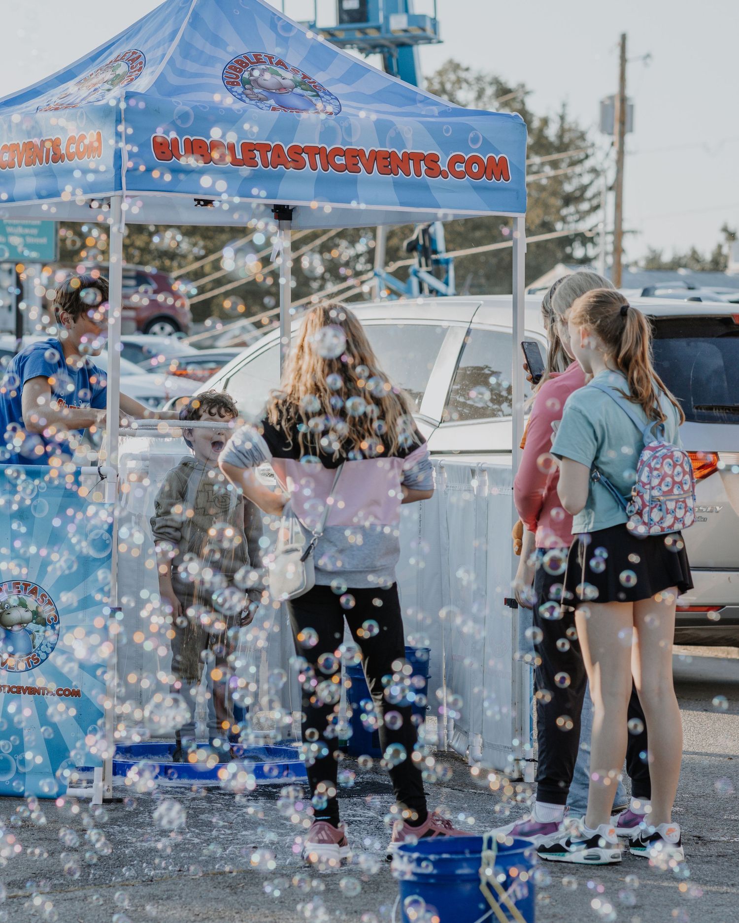 Bubbles floating through the air at an outdoor event with a blue tent and parked cars.