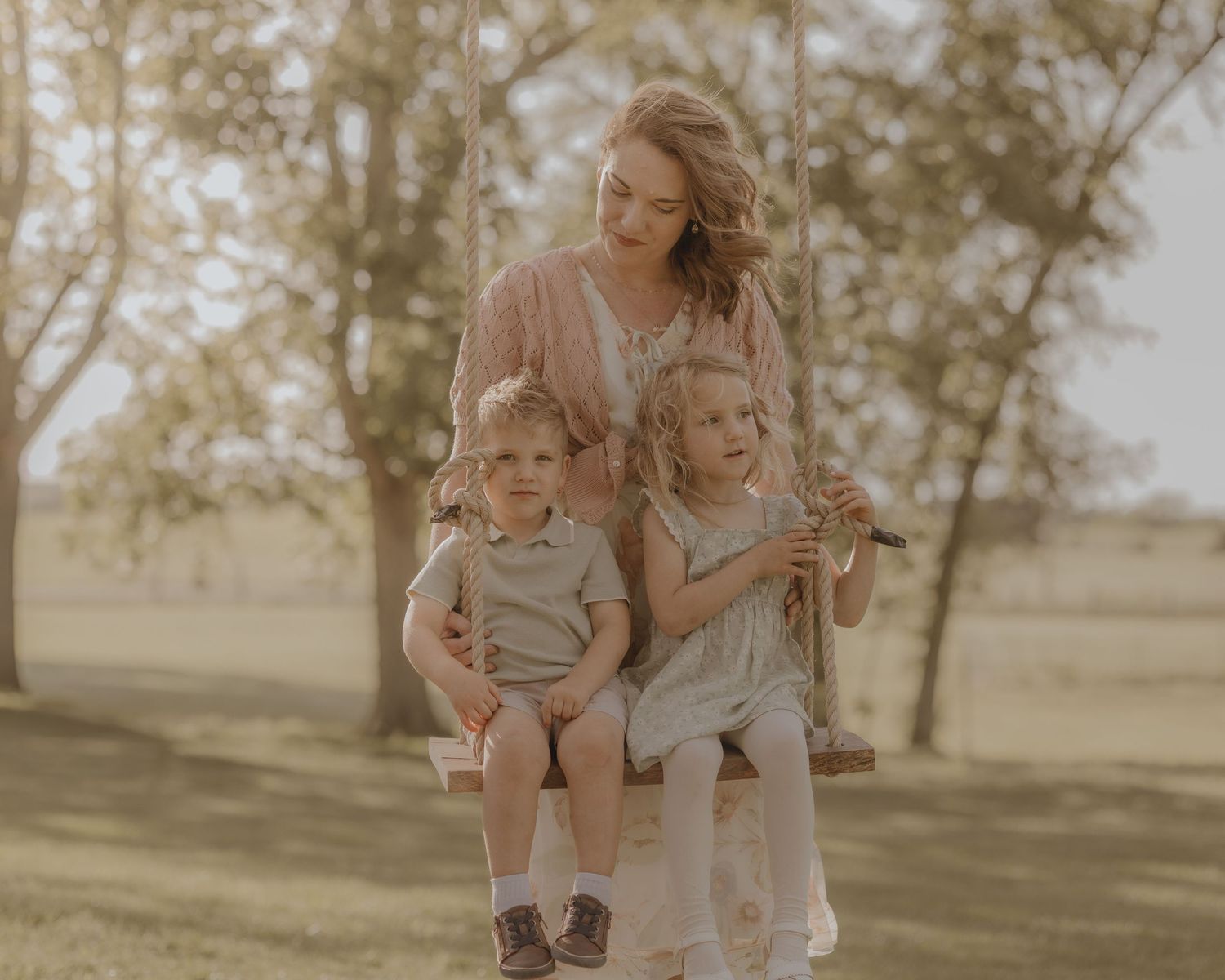 A mother in a floral dress sits on a wooden swing with her two young children in a park.