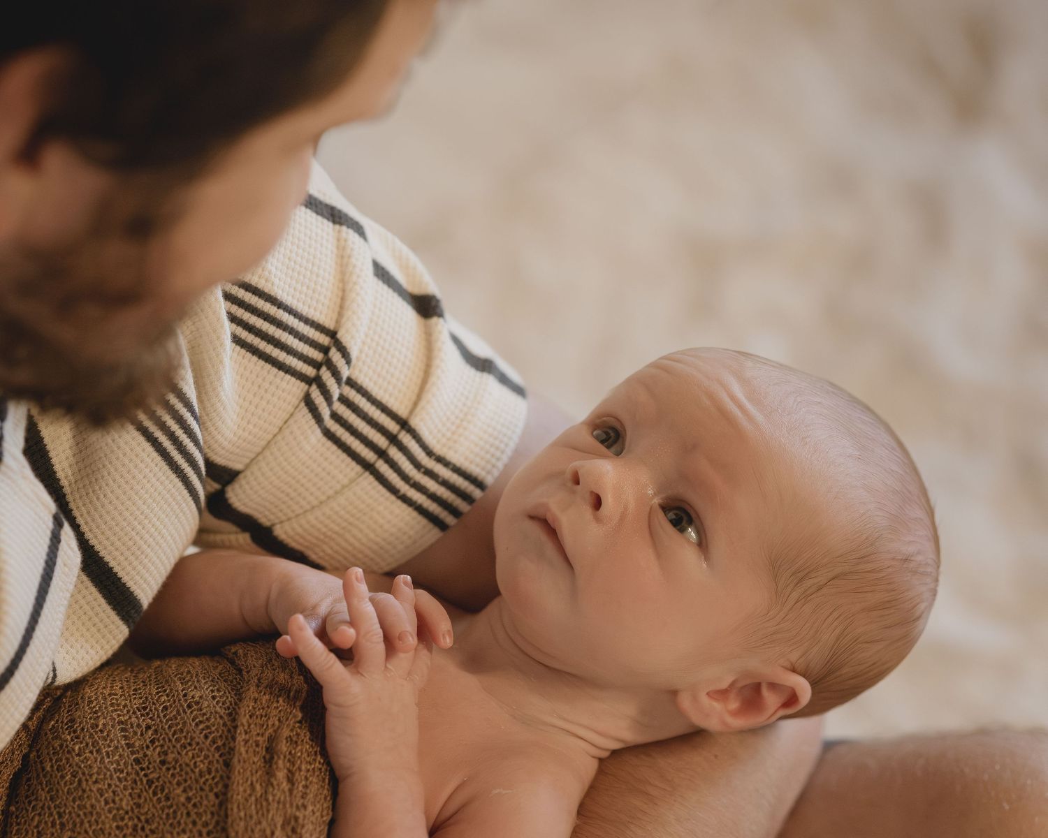 Father in a striped shirt gazing down at his alert newborn baby on a soft rug.