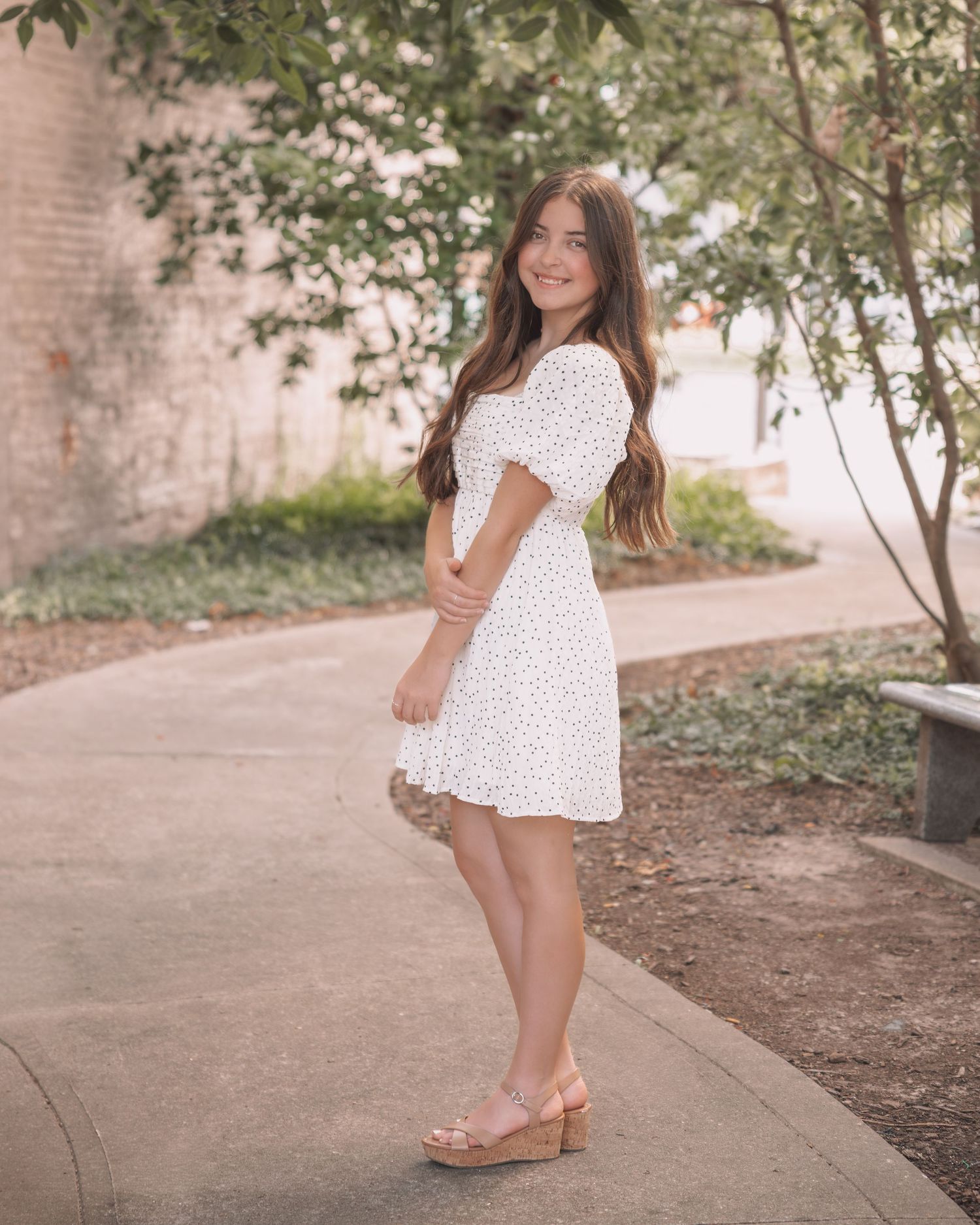 Young woman in white floral mini dress and wedge sandals posing on a pathway surrounded by lush green trees.