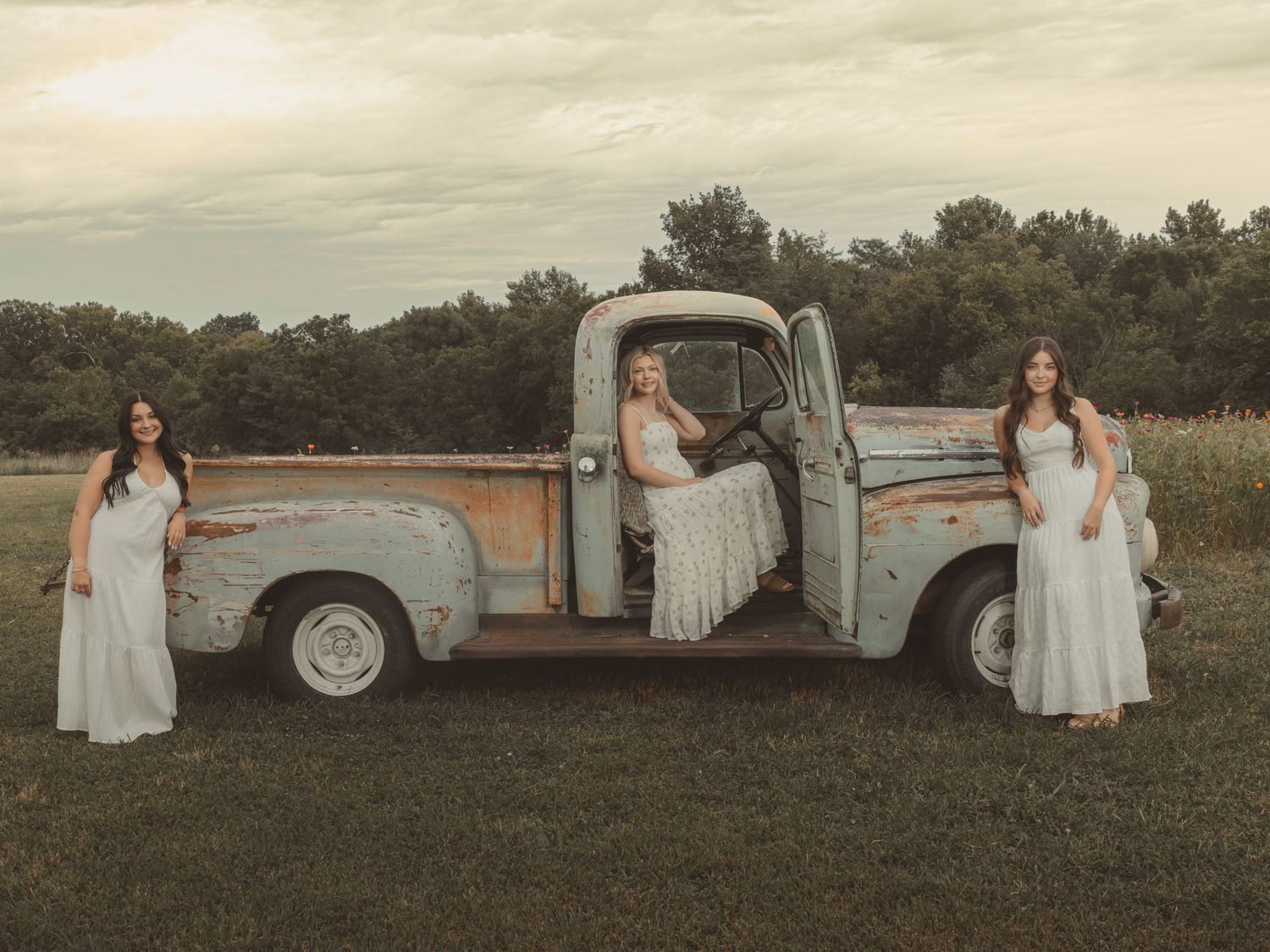Three women in white dresses pose with a vintage rustic truck in a green field at dusk.