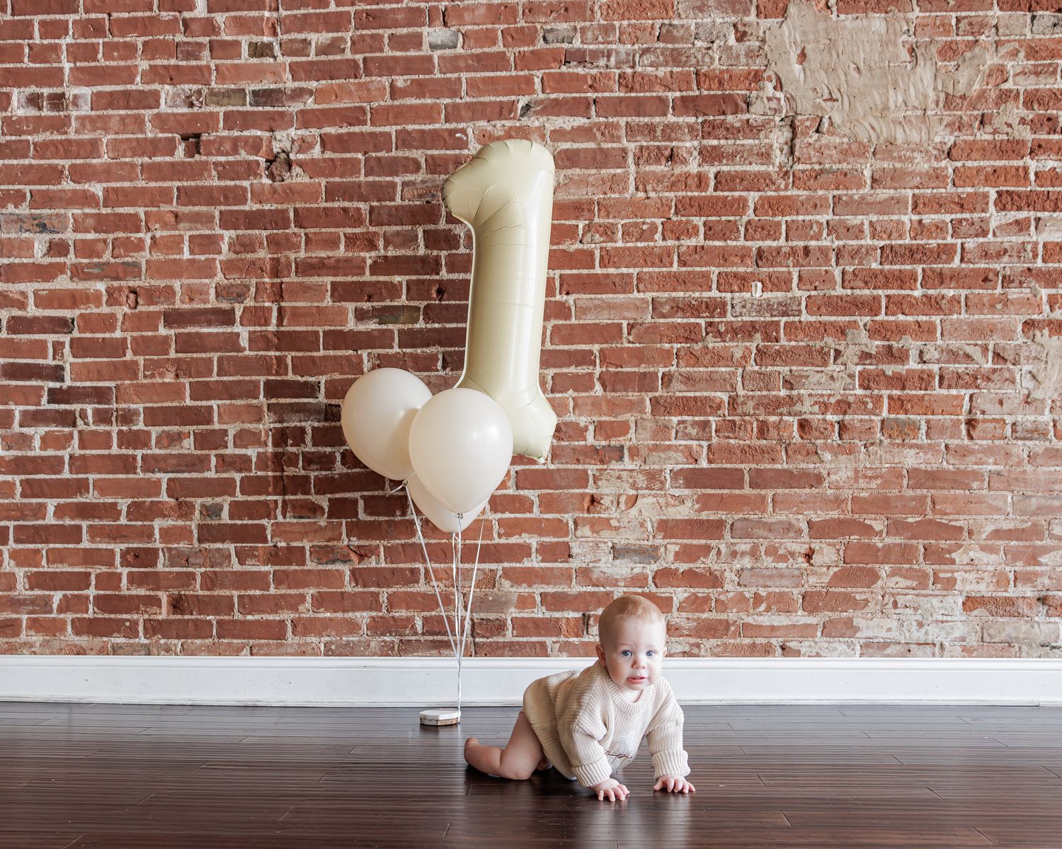 Baby crawling on wood floor beside number one balloon bouquet against rustic brick wall for first birthday.
