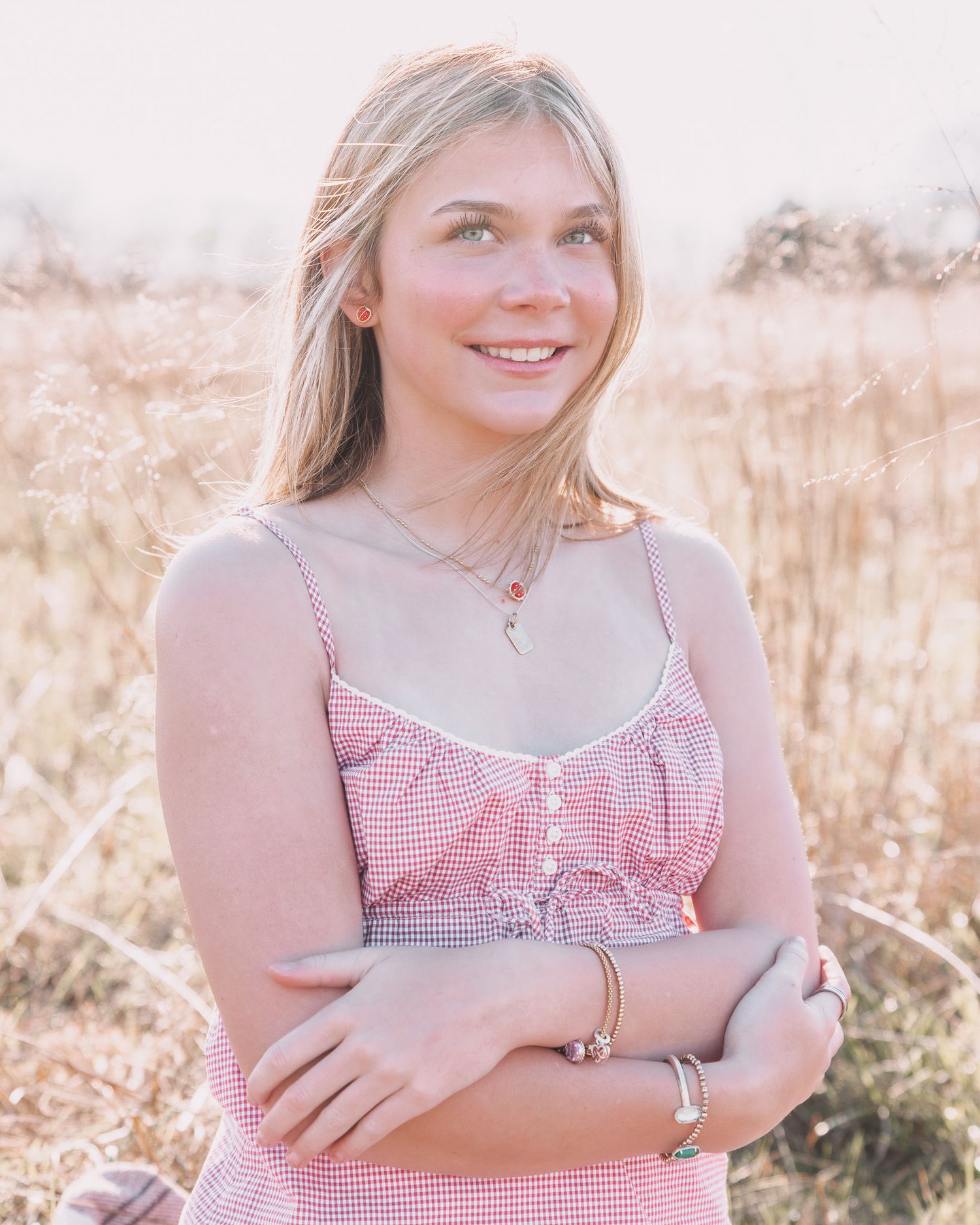 Young woman in red gingham dress smiling outdoors in golden field, wearing delicate necklace and bracelets.