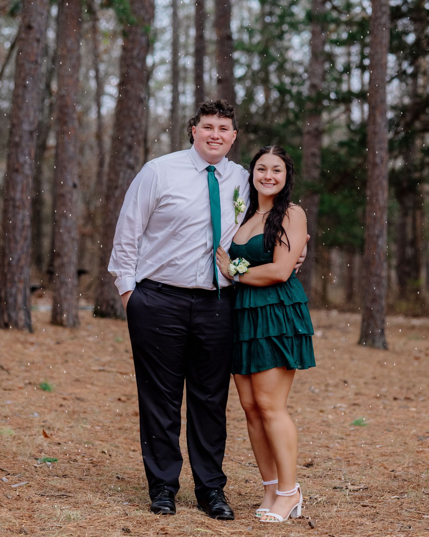 A couple in formal attire poses together in a wooded area, wearing teal and white outfits for photos.