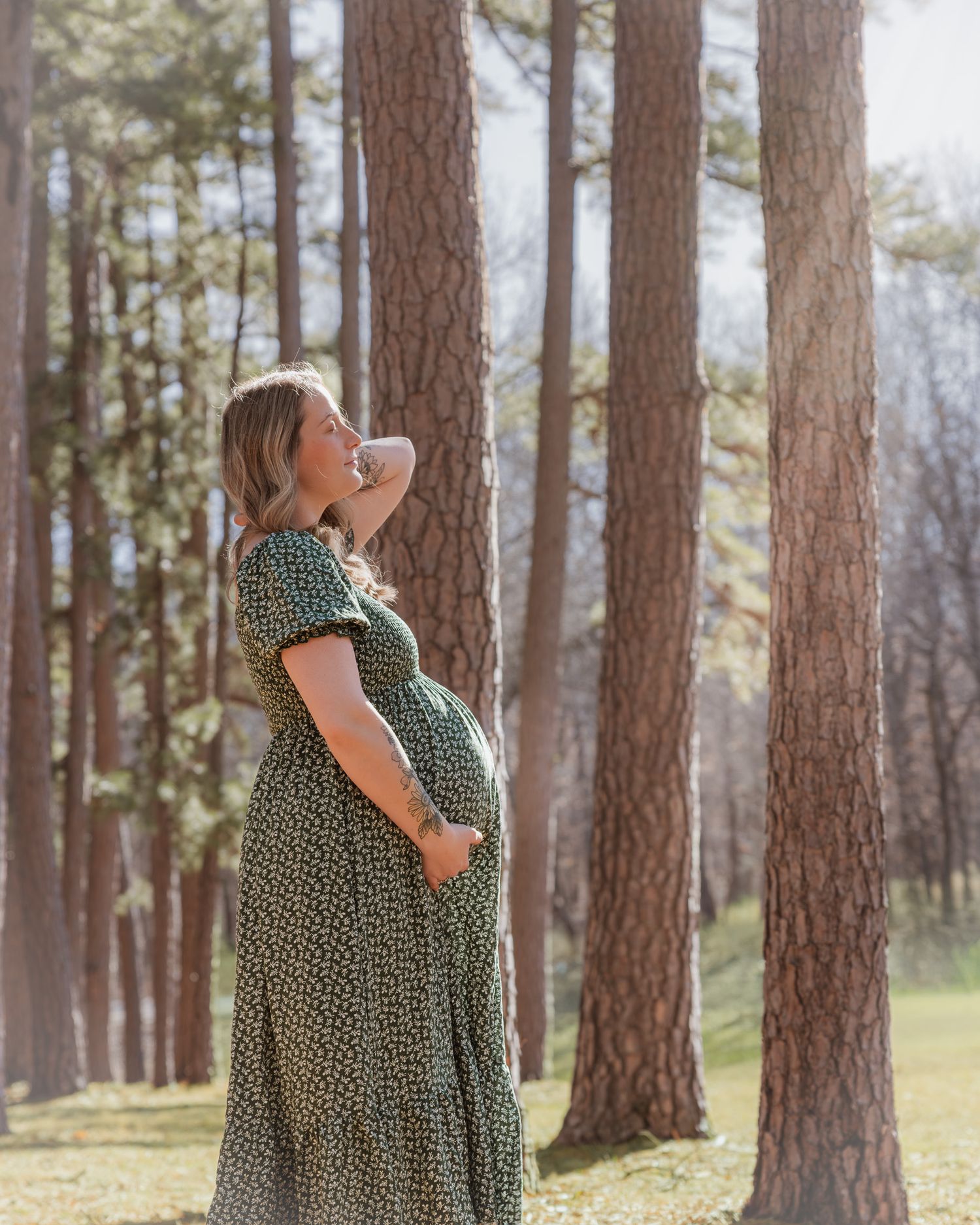 Pregnant woman in floral maxi dress standing among tall pine trees, smiling serenely in a sunlit forest.