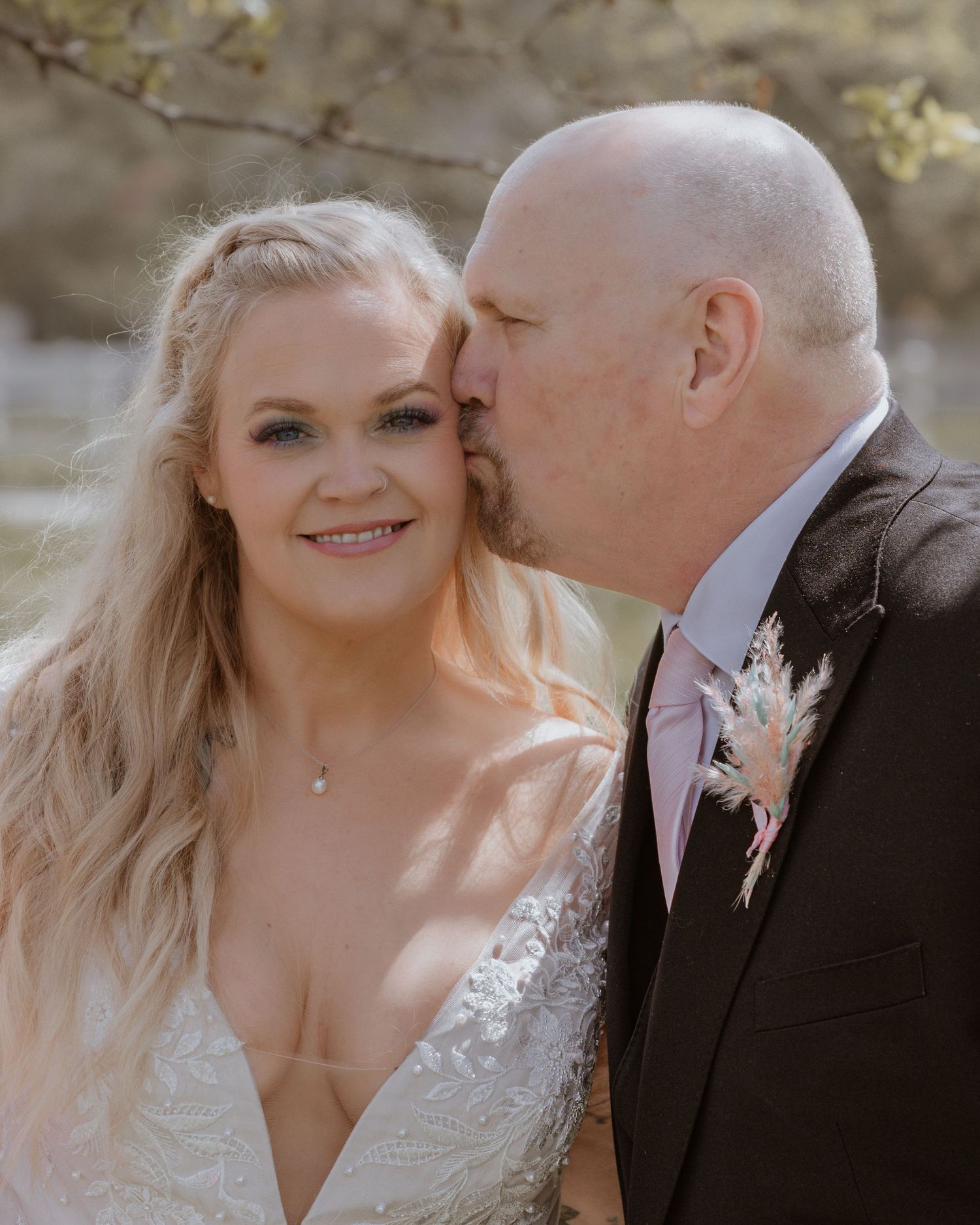 A groom in a dark suit kisses the cheek of a smiling bride in a lace wedding dress outdoors.