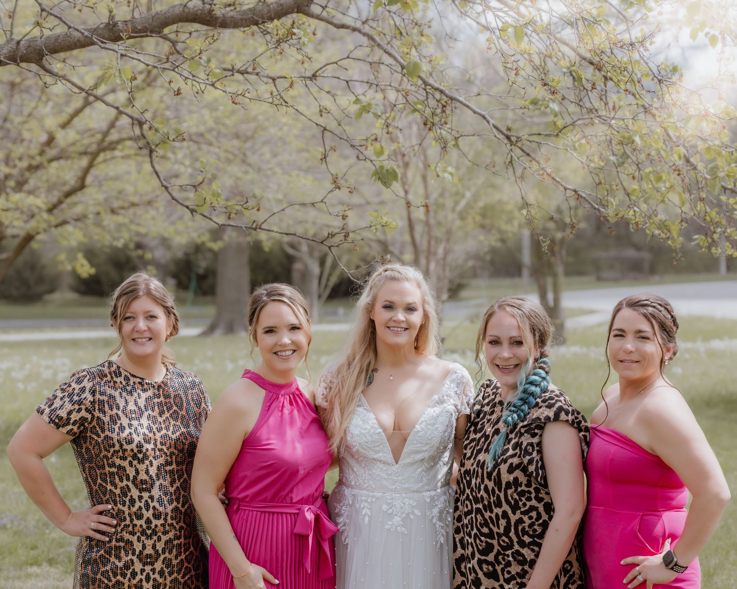 Bride in white wedding gown posing with four bridesmaids in pink and leopard print dresses outdoors.