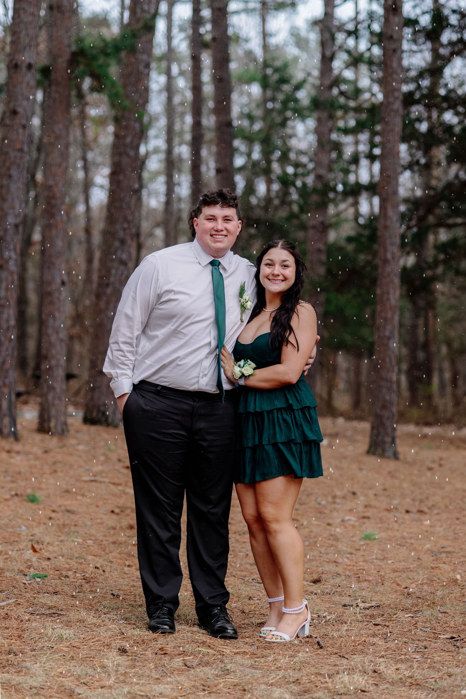 A couple in formal attire poses together in a forest setting, wearing green and white coordinated outfits.