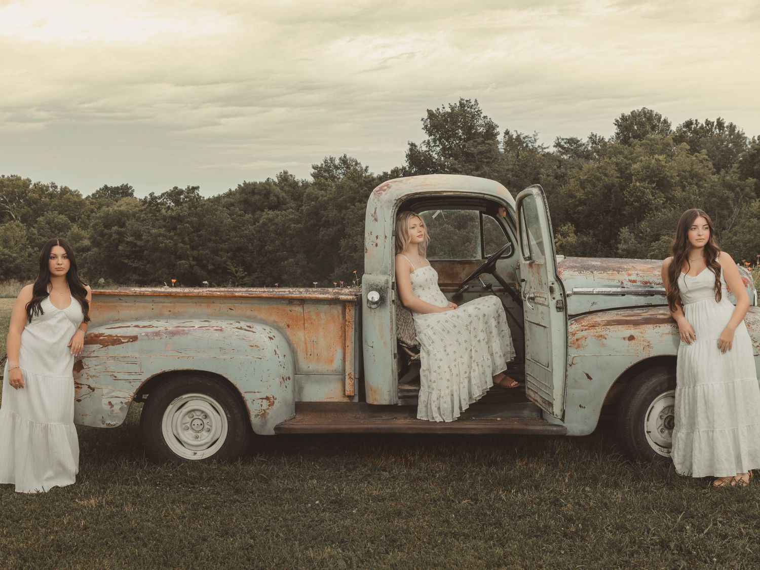 Three women in white dresses pose with a vintage rusty pickup truck in a green field.