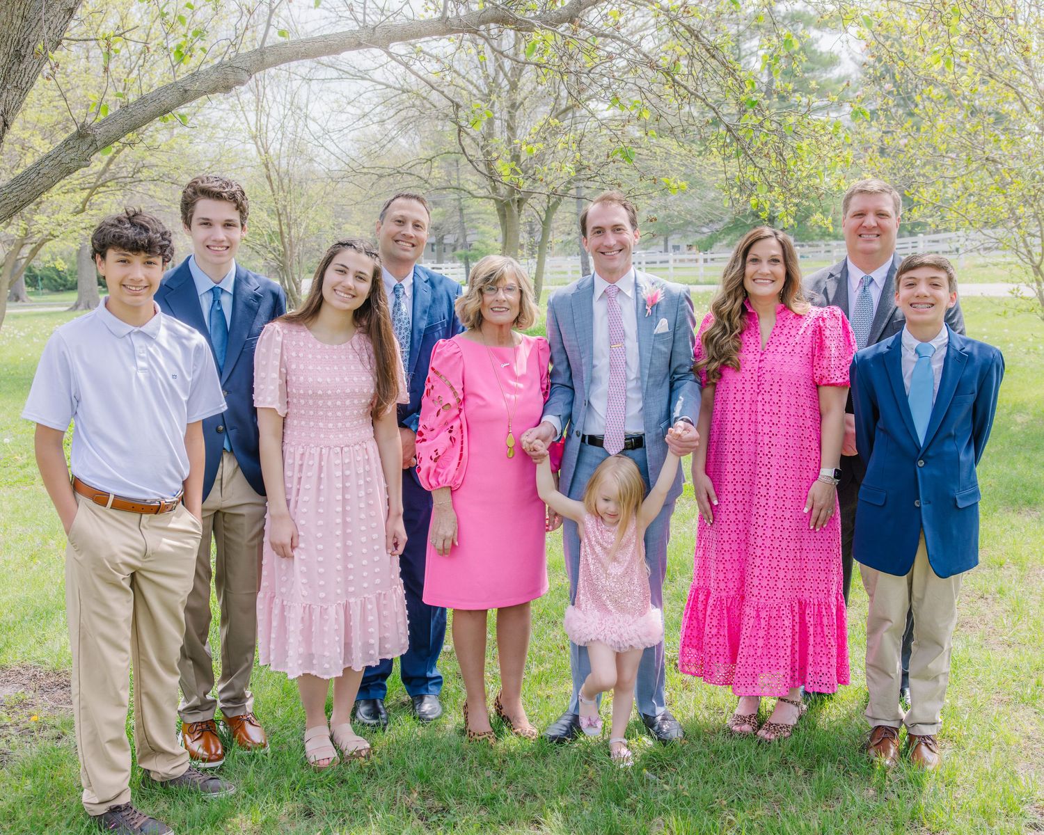 Large family group smiling outdoors in spring, wearing pink and blue formal attire.