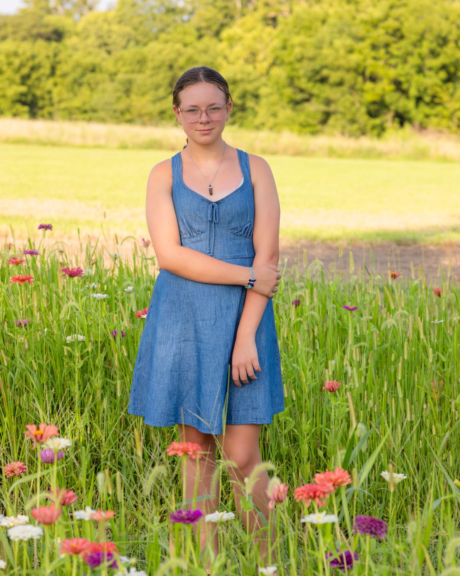 Young woman in blue denim dress standing in colorful wildflower field with green trees in background.