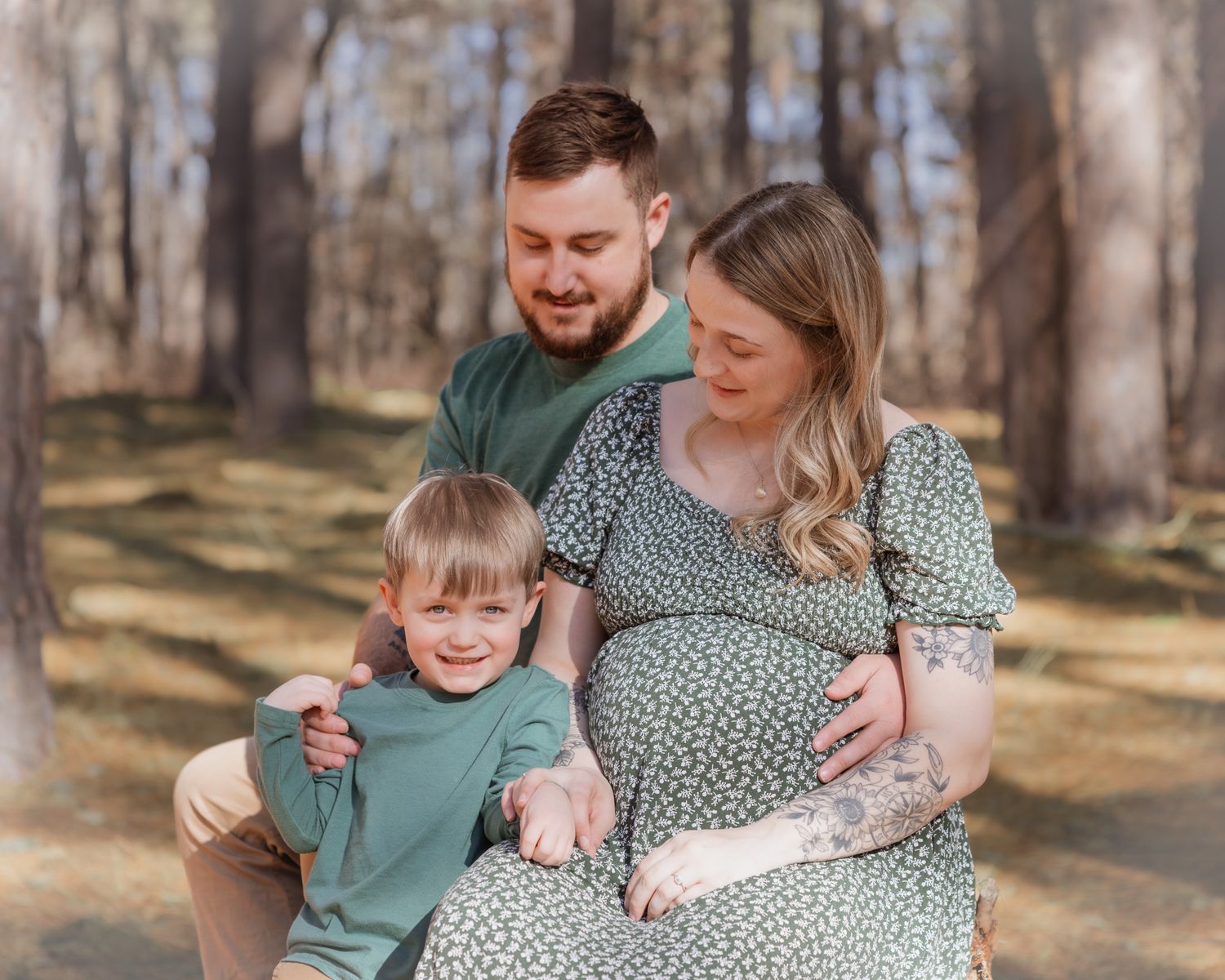 Expecting family maternity photo in forest, parents smiling with young boy in matching green outfits.