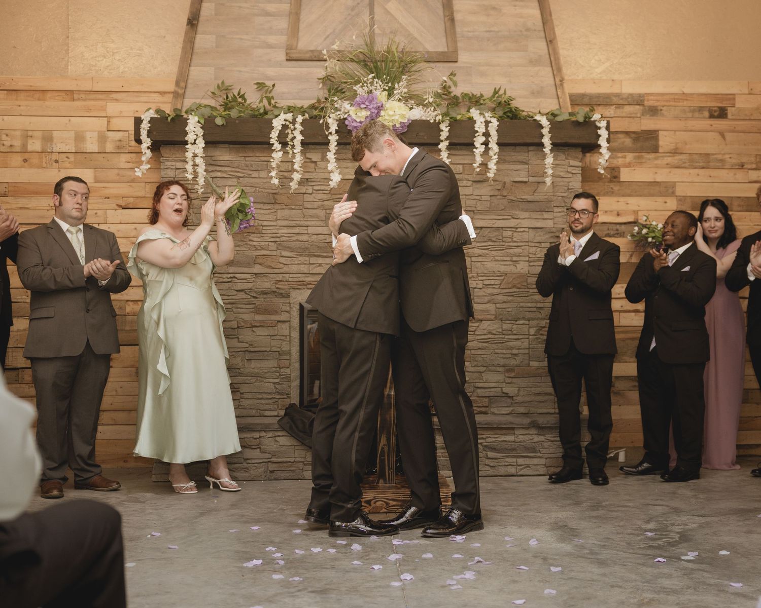 Two grooms share a first kiss at their rustic barn wedding ceremony while guests applaud.