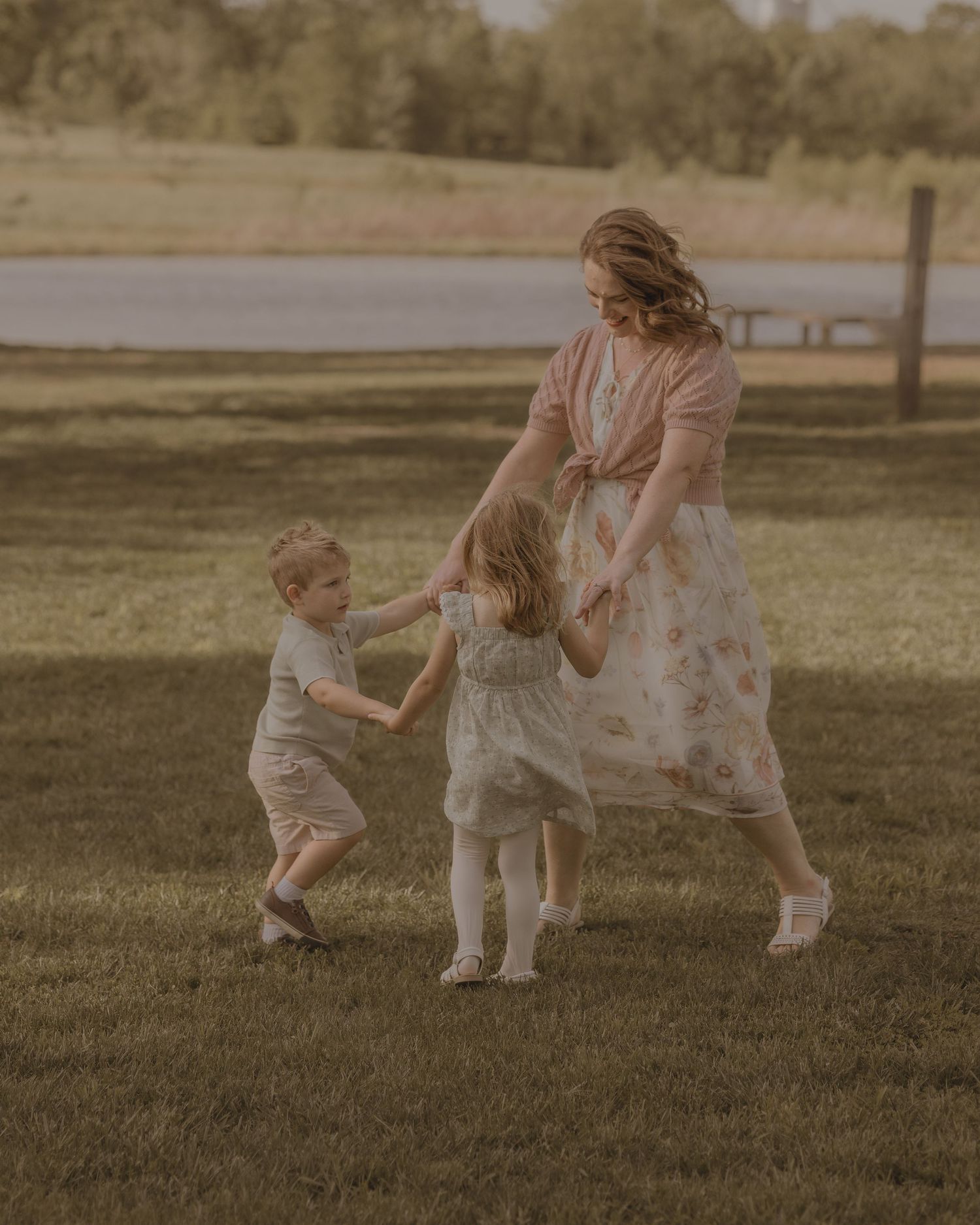 Mother playfully dancing with two young children on green grass during golden hour outdoor family photo session.