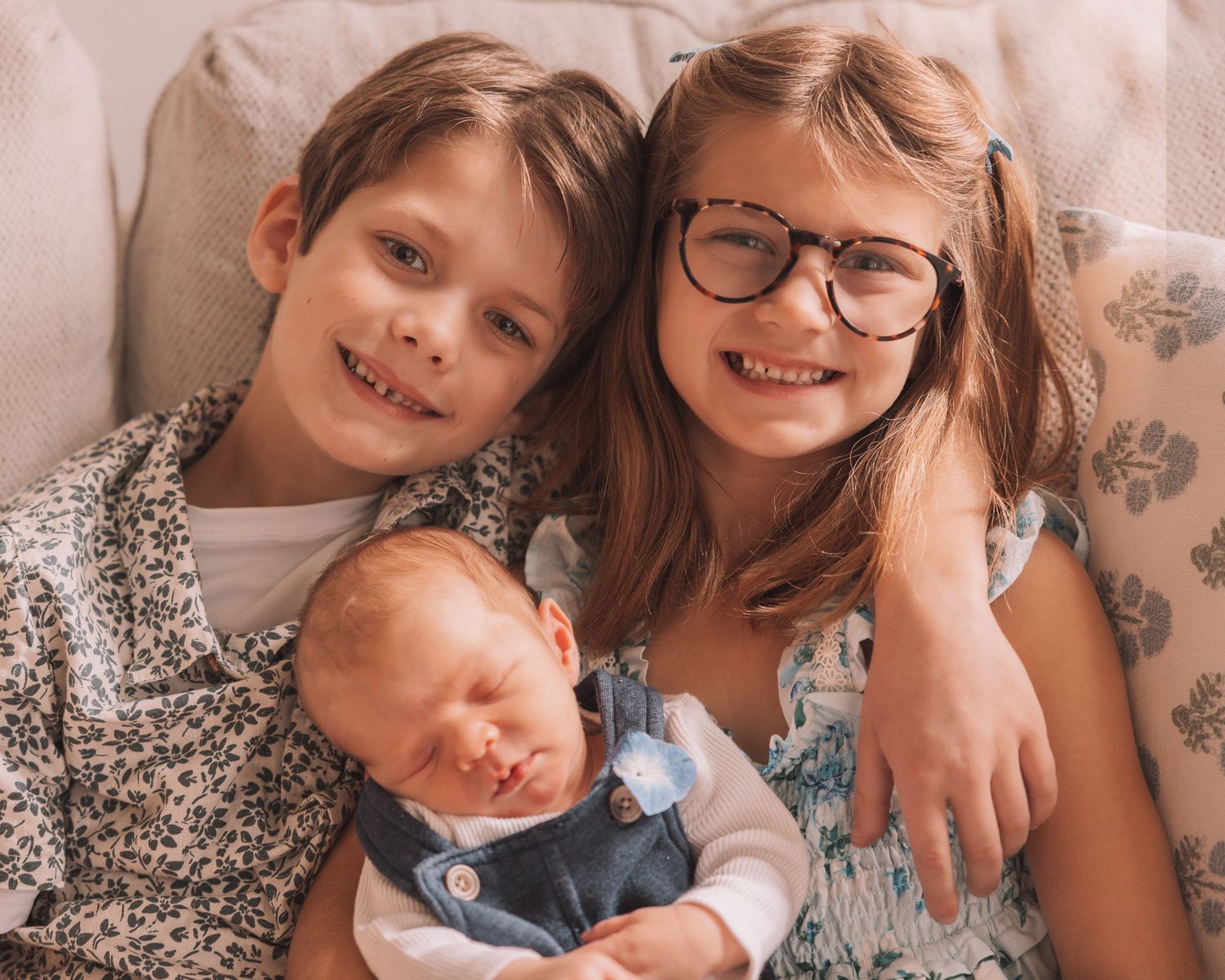 Two smiling children pose with a sleeping newborn baby on a cozy couch.