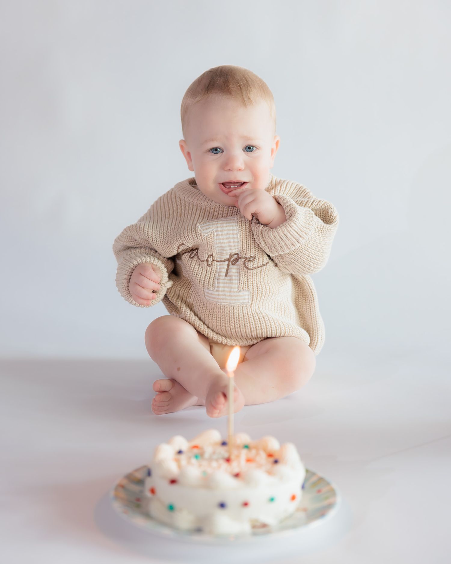 Baby in beige knit romper sits before a white birthday cake with one lit candle during first birthday smash cake photo.