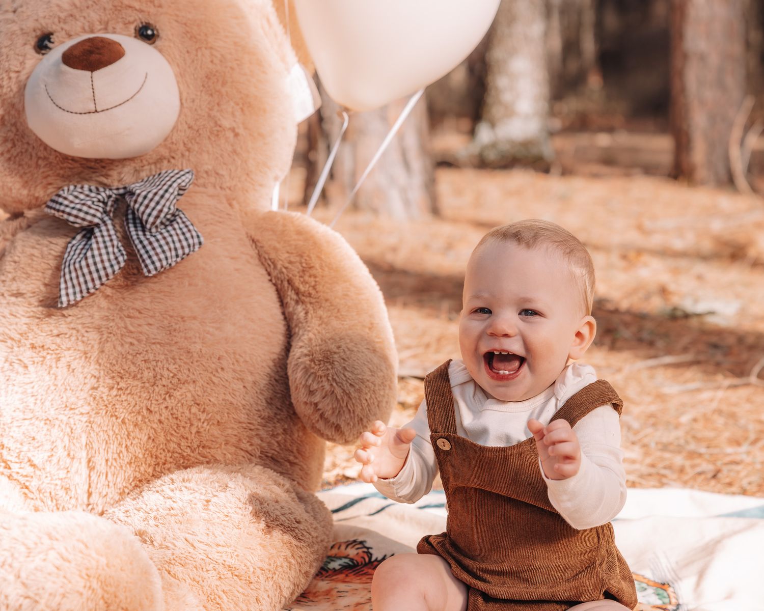 Baby sits next to large teddy bear in autumn forest setting with warm lighting.
