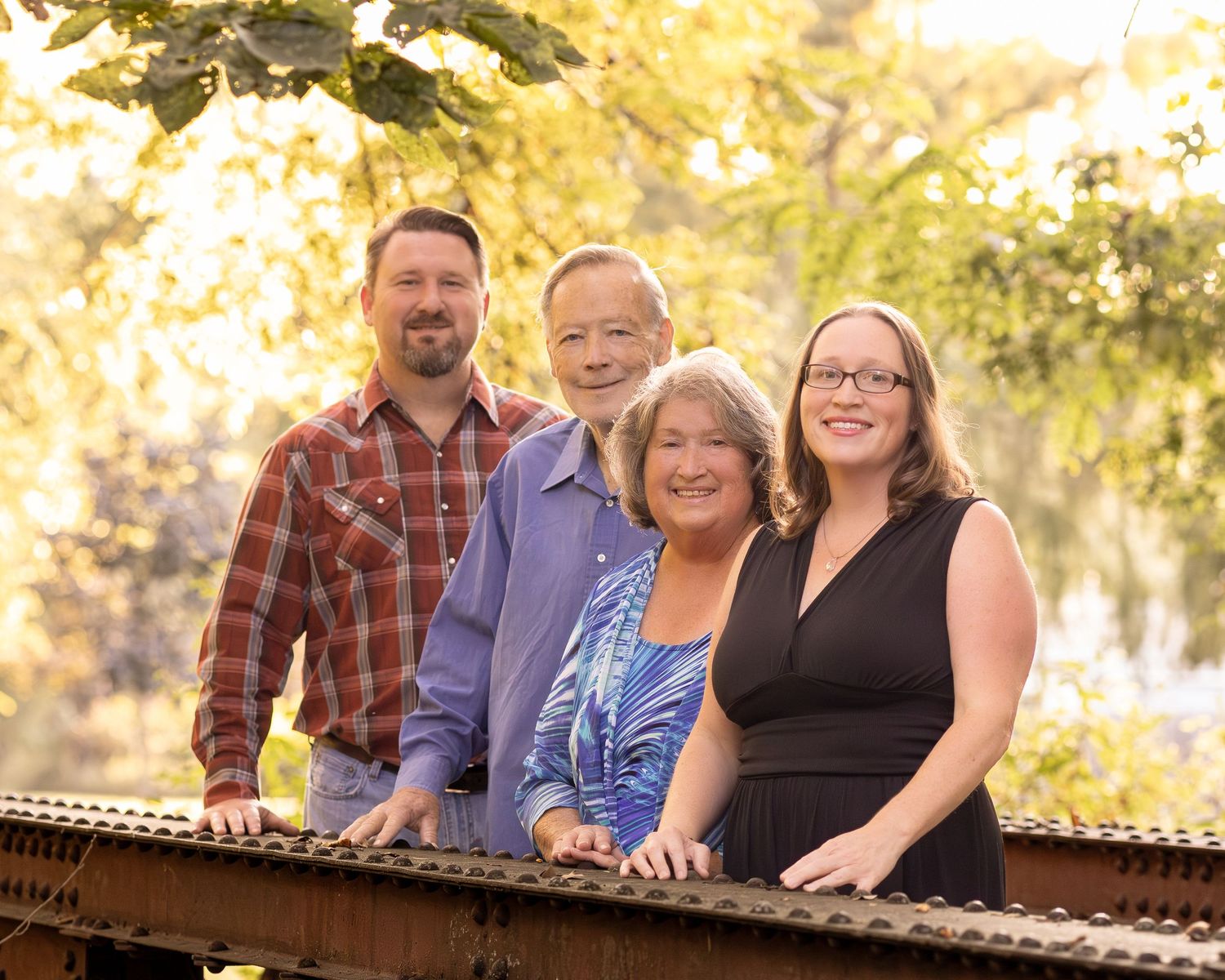 Four smiling adults pose outdoors on a bridge surrounded by lush green and golden foliage in a family portrait.
