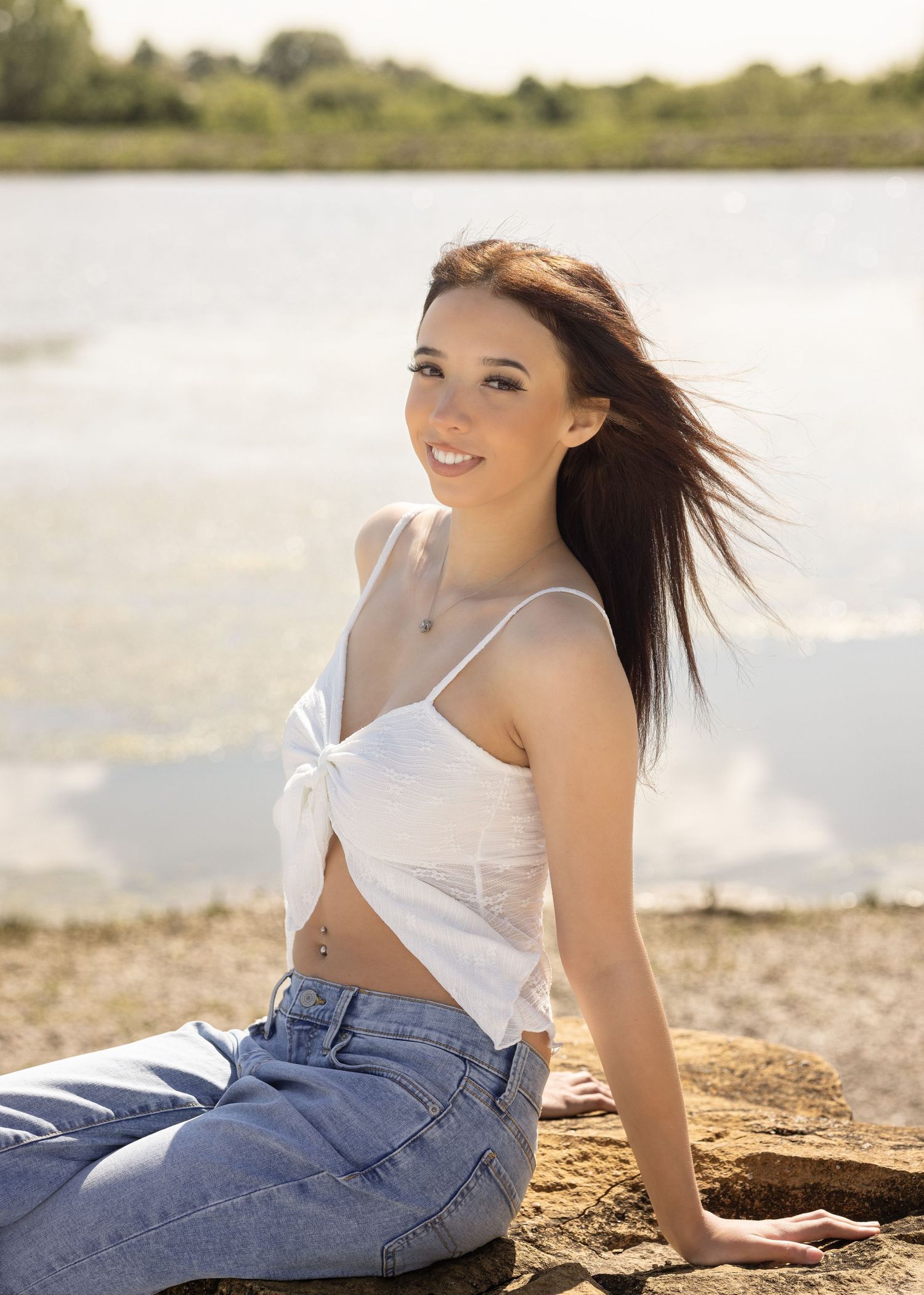Young woman with dark hair in white crop top and jeans sitting by a lake shore on a sunny day.