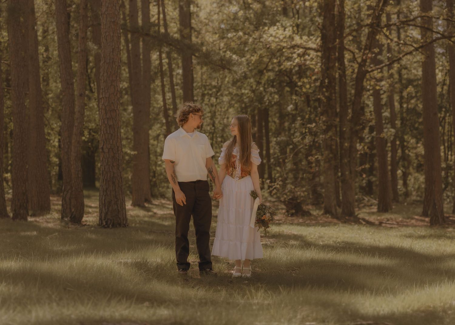 A couple walks hand in hand through a sunlit forest clearing surrounded by tall trees.