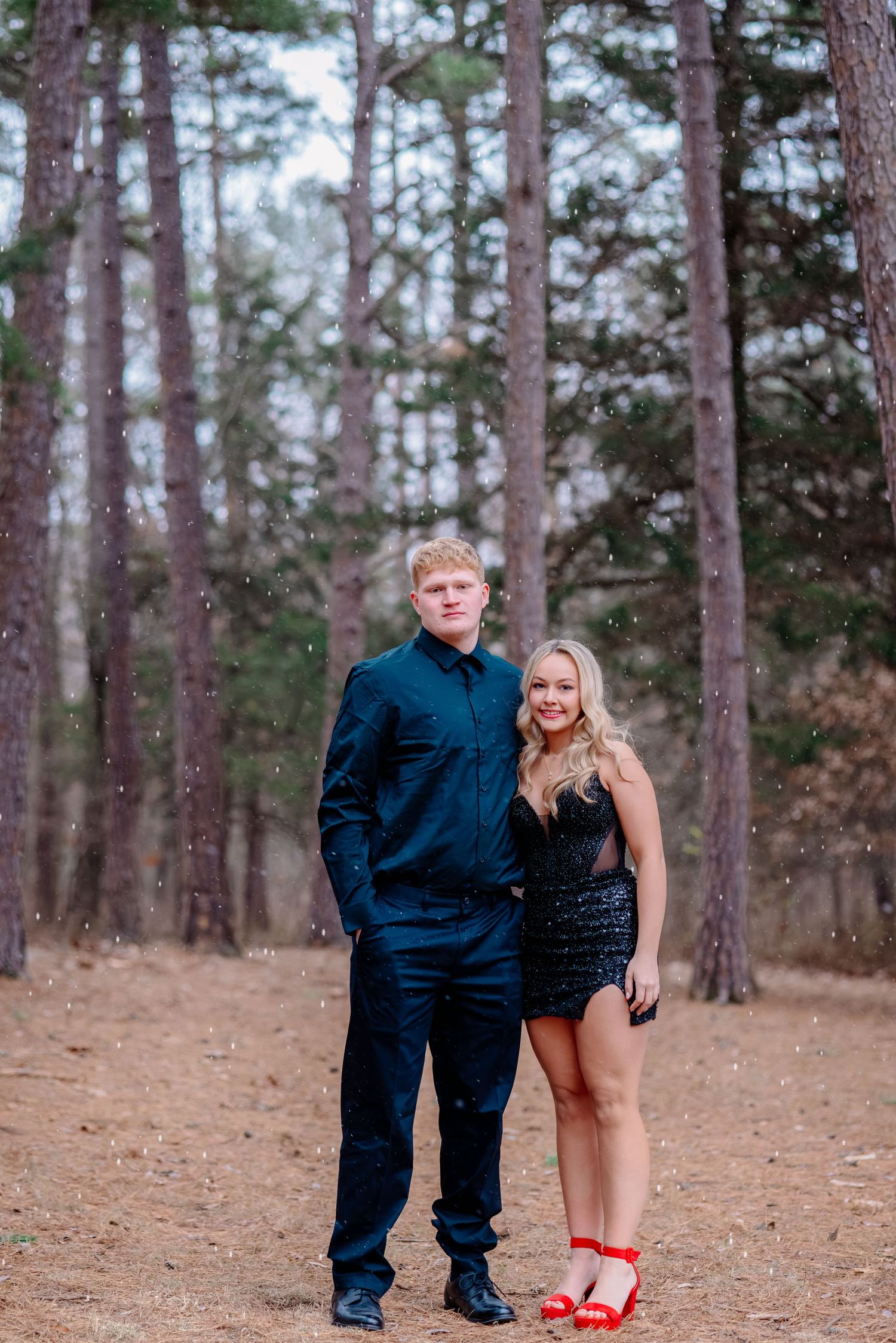 A couple poses together in formal attire against a backdrop of tall pine trees on a dirt path.