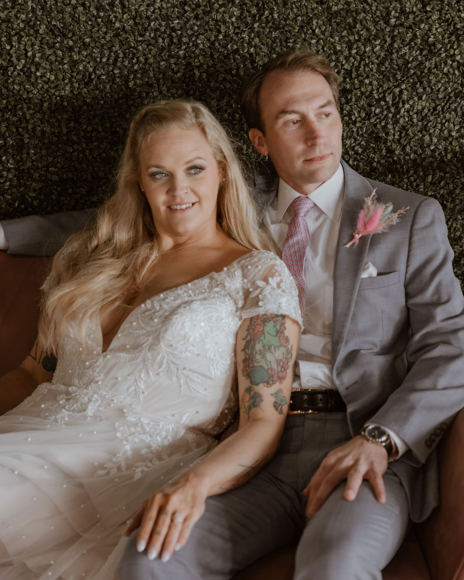 Bride in lace wedding gown and groom in gray suit with pink boutonniere sitting together at wedding reception.