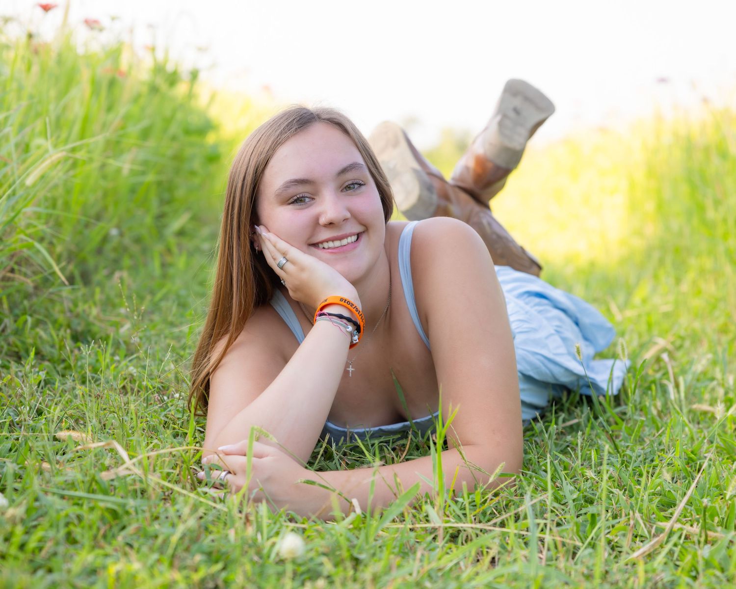 Smiling teenage girl lying on green grass in a sunny field, wearing a light blue top and orange bracelet.